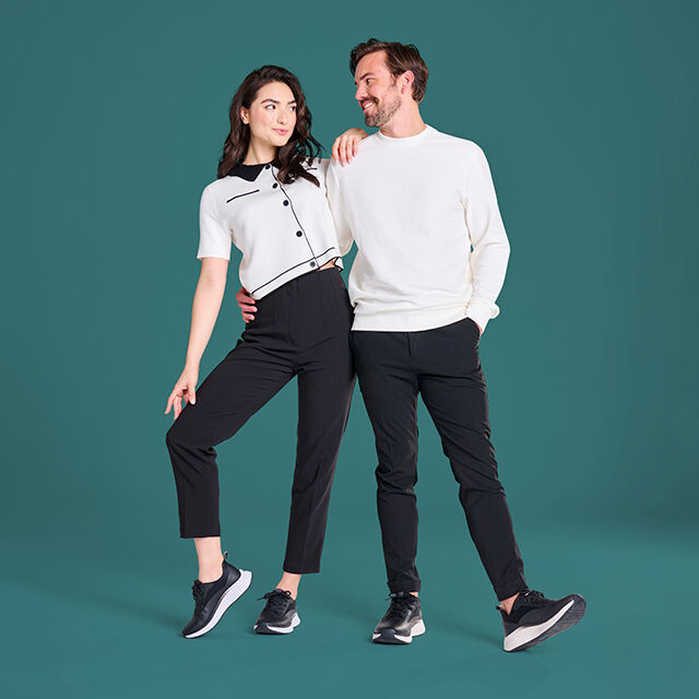 Man and Woman, both wearing white tops and black bottoms, and the same style of sneaker, Tiberia in Black for her, Tacoda in Black for him, each with one shoe in profile